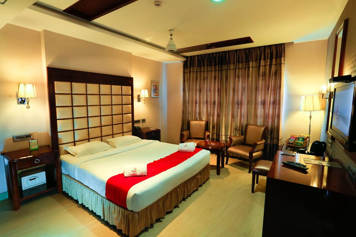 A hotel room with a large bed, a desk with a television, a chair, and a desk lamp.