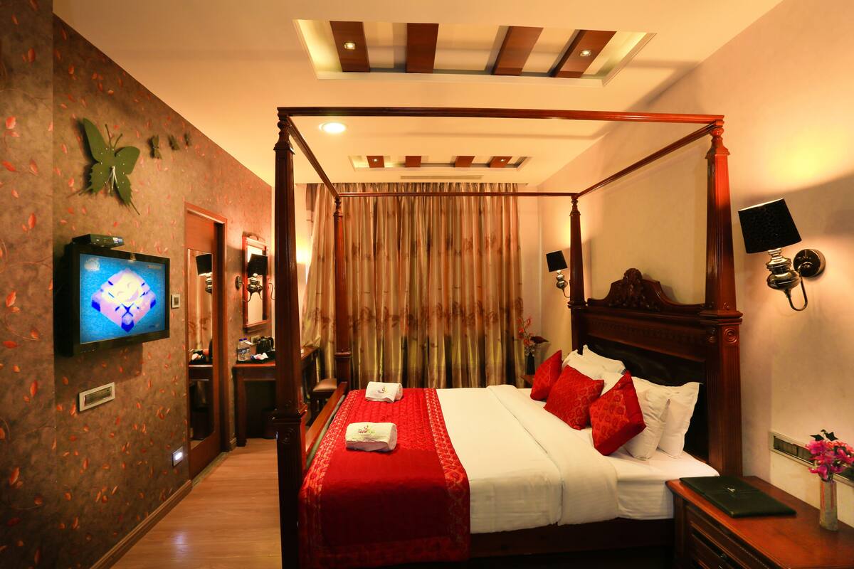 A four-poster bed with red and white linens, a television on the left wall, and a wooden nightstand with a lamp and a vase of flowers.