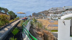 A coastal view with a beach, a pathway with greenery, and a building with a white facade. - Apt in Puerto Rico 100m From the Beach, Free Wifi, Community Pool and Garden (Las Palmas)