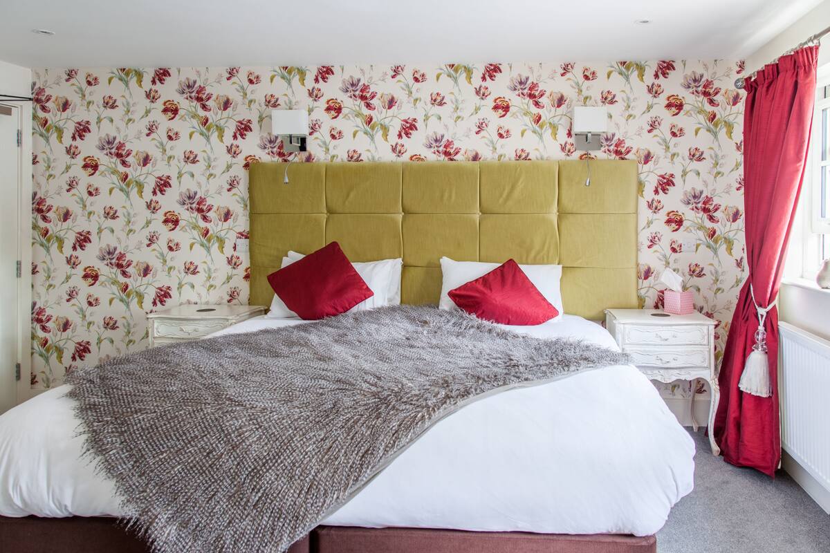 A bedroom with floral wallpaper, a tufted headboard, a bed with white and red bedding, a grey throw, and a nightstand with a lamp.