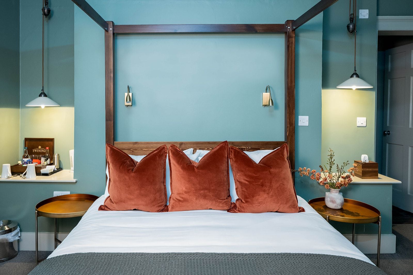 Quarto de hotel com cama, duas mesas de cabeceira, um criado-mudo, luminária na parede e um vaso com flores.