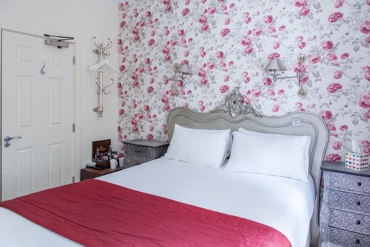 A neatly made bed with a red blanket, a white door, a nightstand with a lamp, and a dresser with a patterned drawer.