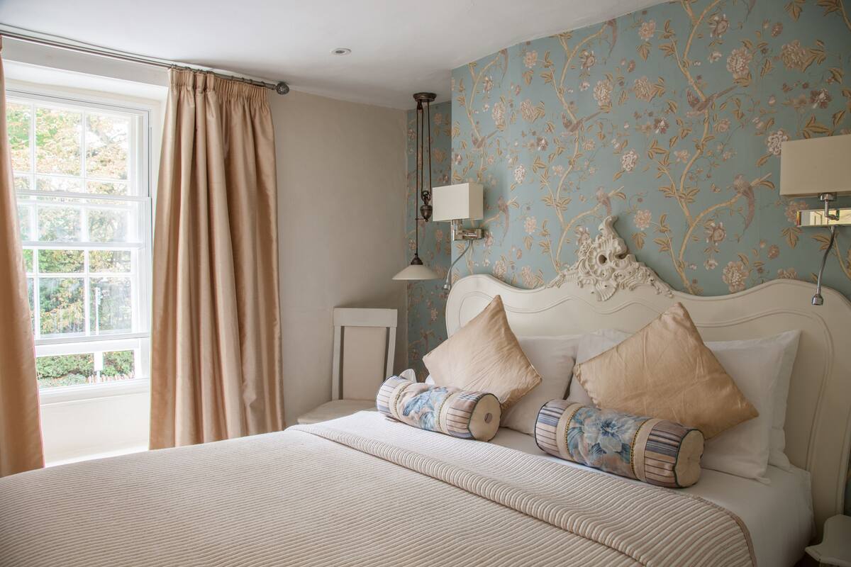 A bedroom with a large bed, floral wallpaper, and a window with curtains.