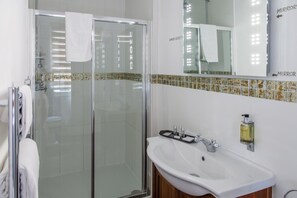 A modern bathroom with a glass shower enclosure, a white sink with a soap dispenser, and a mirror with lights. - The Wychwood Inn (Chipping Norton)