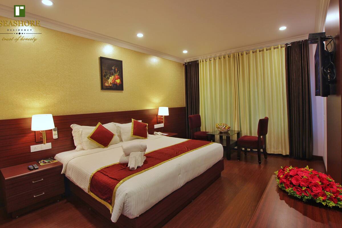 A hotel room with a large bed, two bedside tables, a desk with a chair, a small table with a vase of flowers, and a wall-mounted TV.
