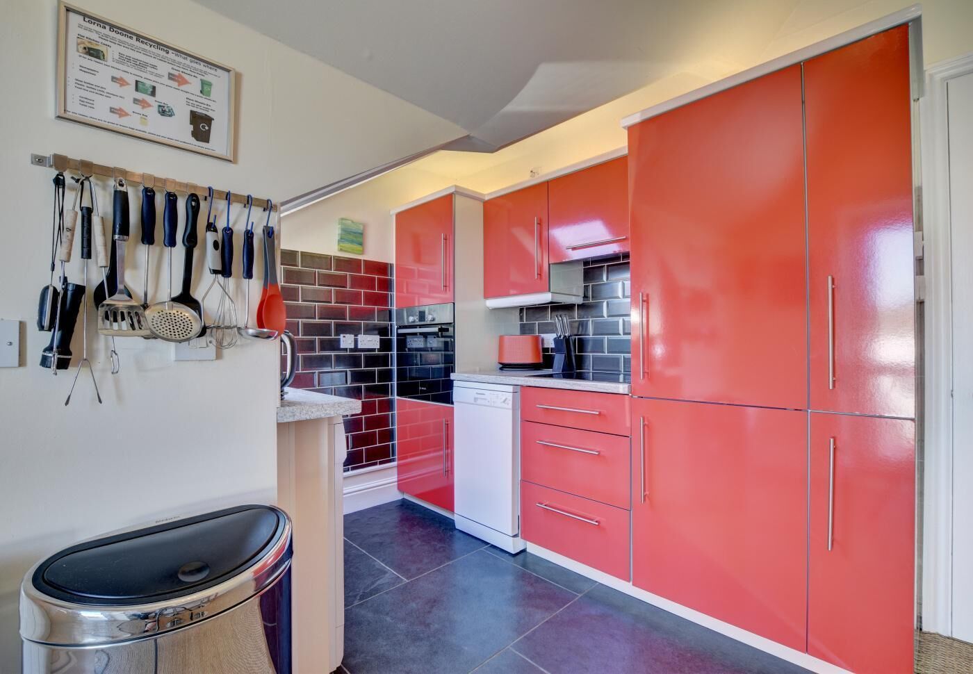 A kitchen with red cabinets, a white dishwasher, and a variety of kitchen utensils hanging on the wall.