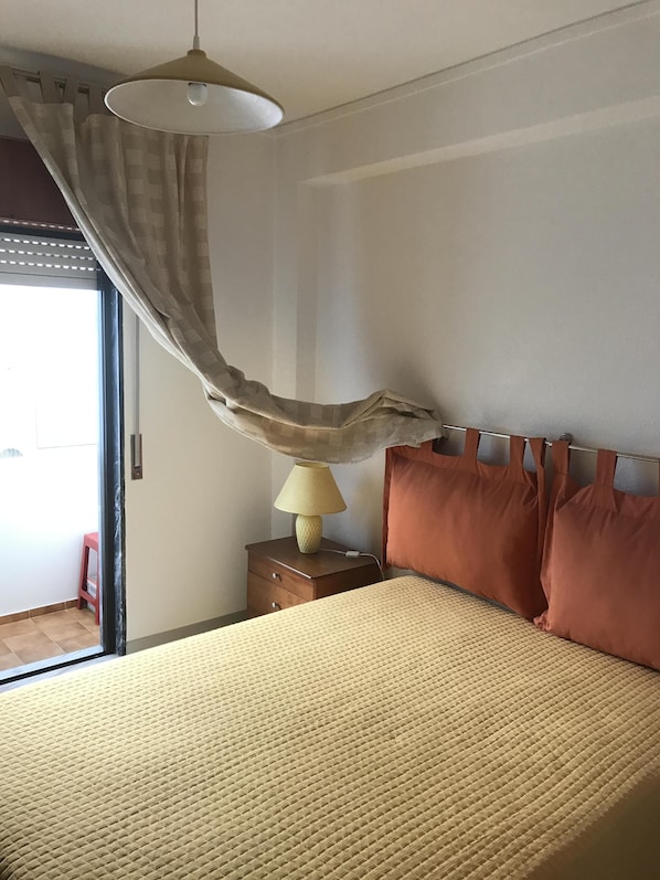 A bedroom with a bed, a nightstand with a lamp, a window with curtains, and a view of the outside.