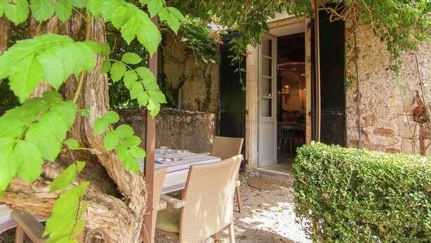 An outdoor dining area with wicker chairs and a table, surrounded by greenery and a stone wall.