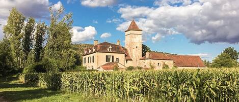 Un grand bâtiment historique, doté d’une tour imposante, entouré d’une végétation luxuriante et d’un champ de maïs.
