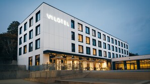 A modern hotel with a prominent 'VILOTEL' sign, multiple floors, and a well-lit entrance with outdoor seating. - VILOTEL - Hotel & Restaurant (Oberkochen)