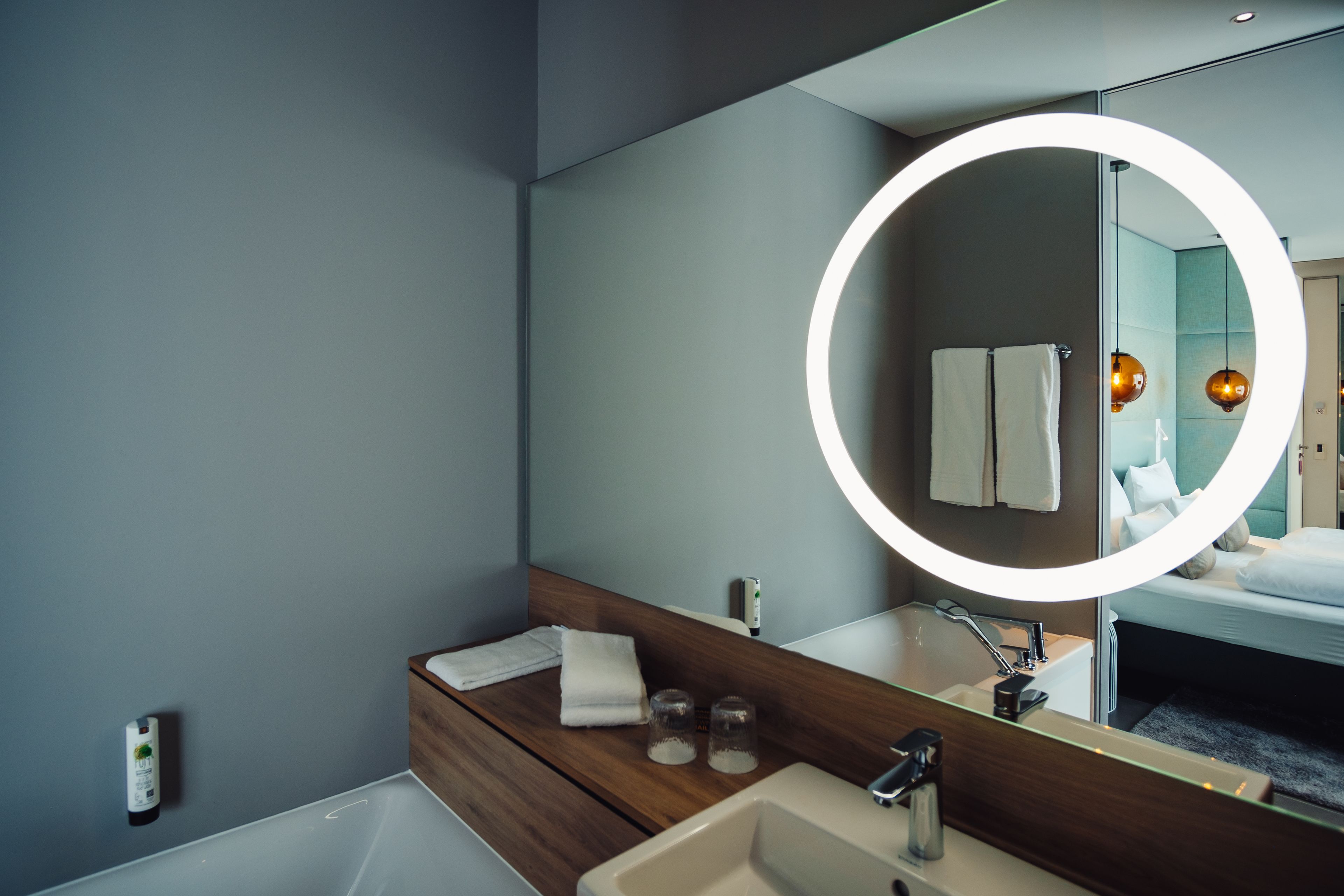 Une salle de bain moderne avec un grand miroir, un meuble-lavabo en bois, une baignoire et un luminaire rond.