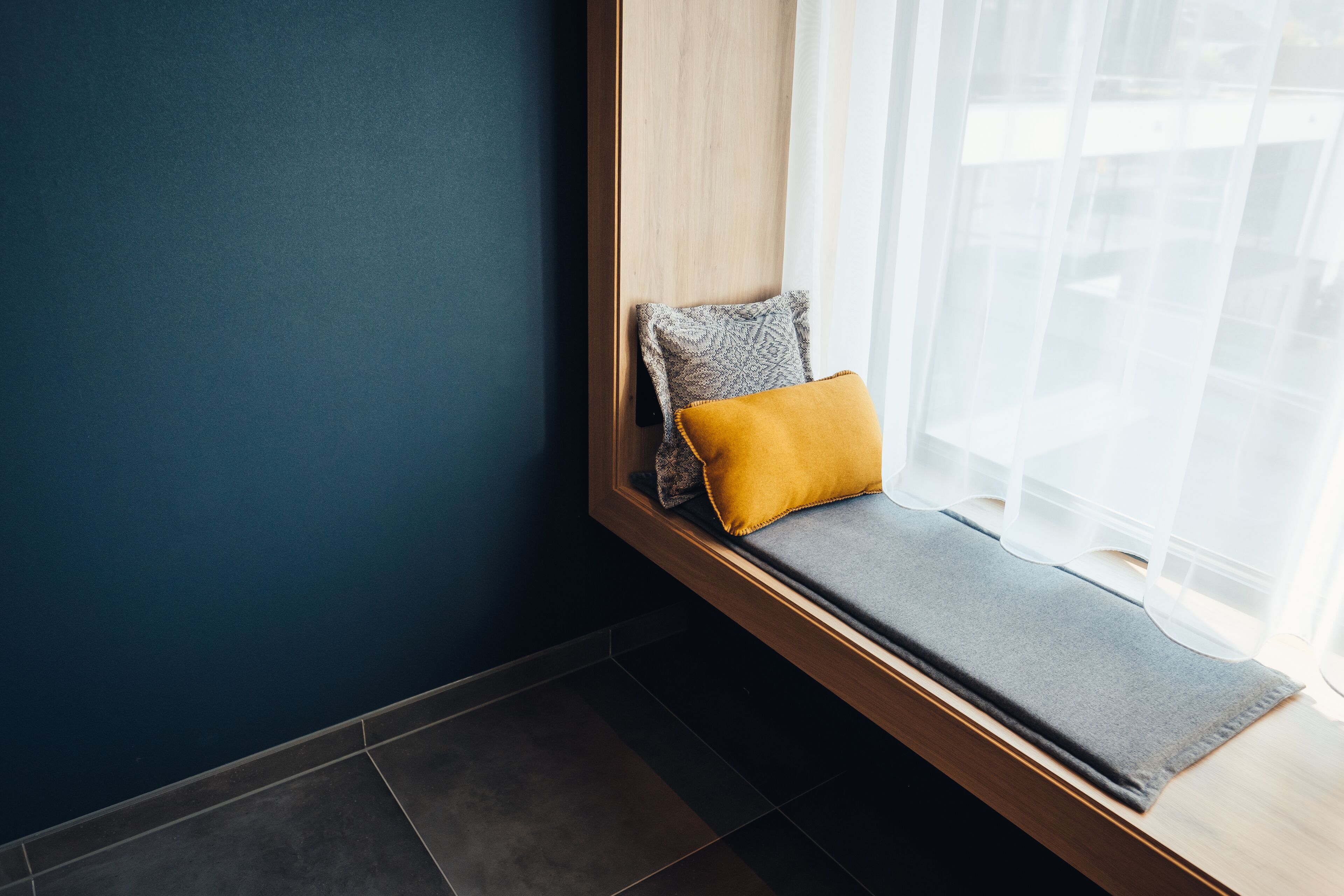 Asiento junto a la ventana en esquina, con cojín gris y almohada amarilla, estructura de madera y vista a un edificio a través de cortinas traslúcidas.