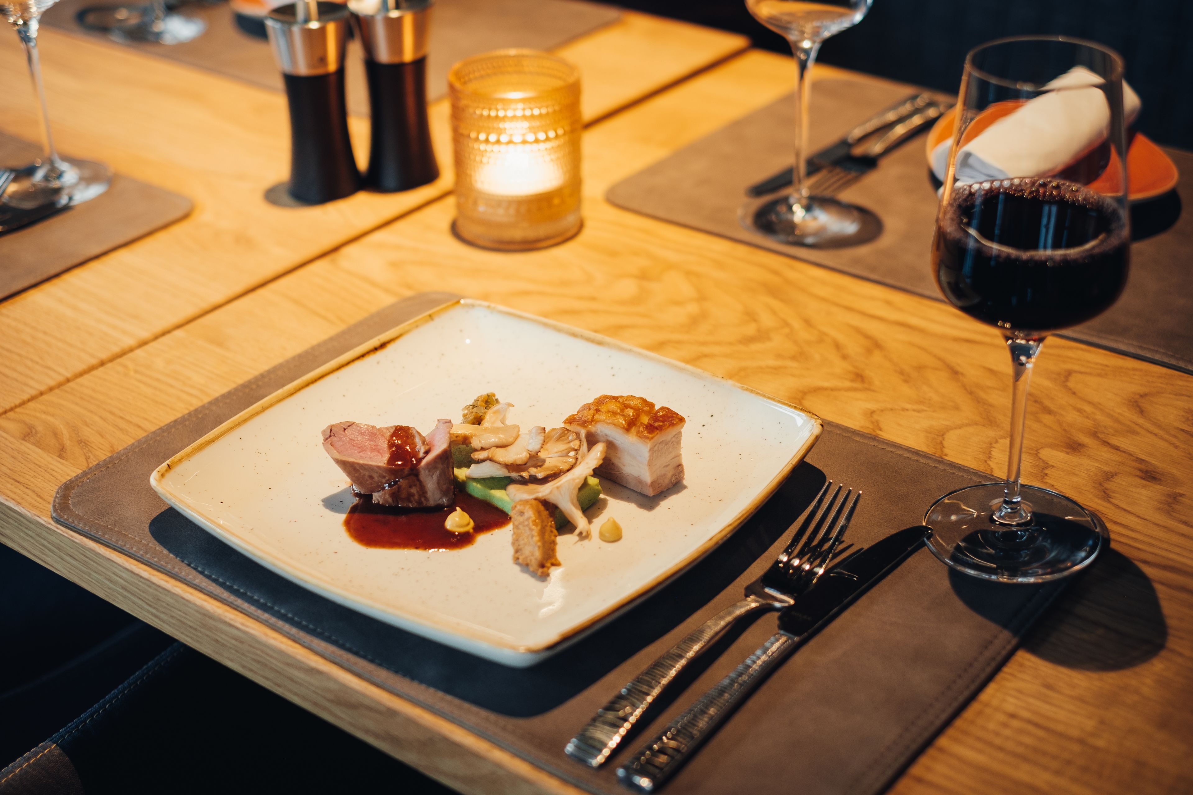 A dining table with a square plate of food, a glass of red wine, and a candle.