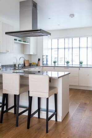 A modern kitchen with a central island, bar stools, and a window with blinds.