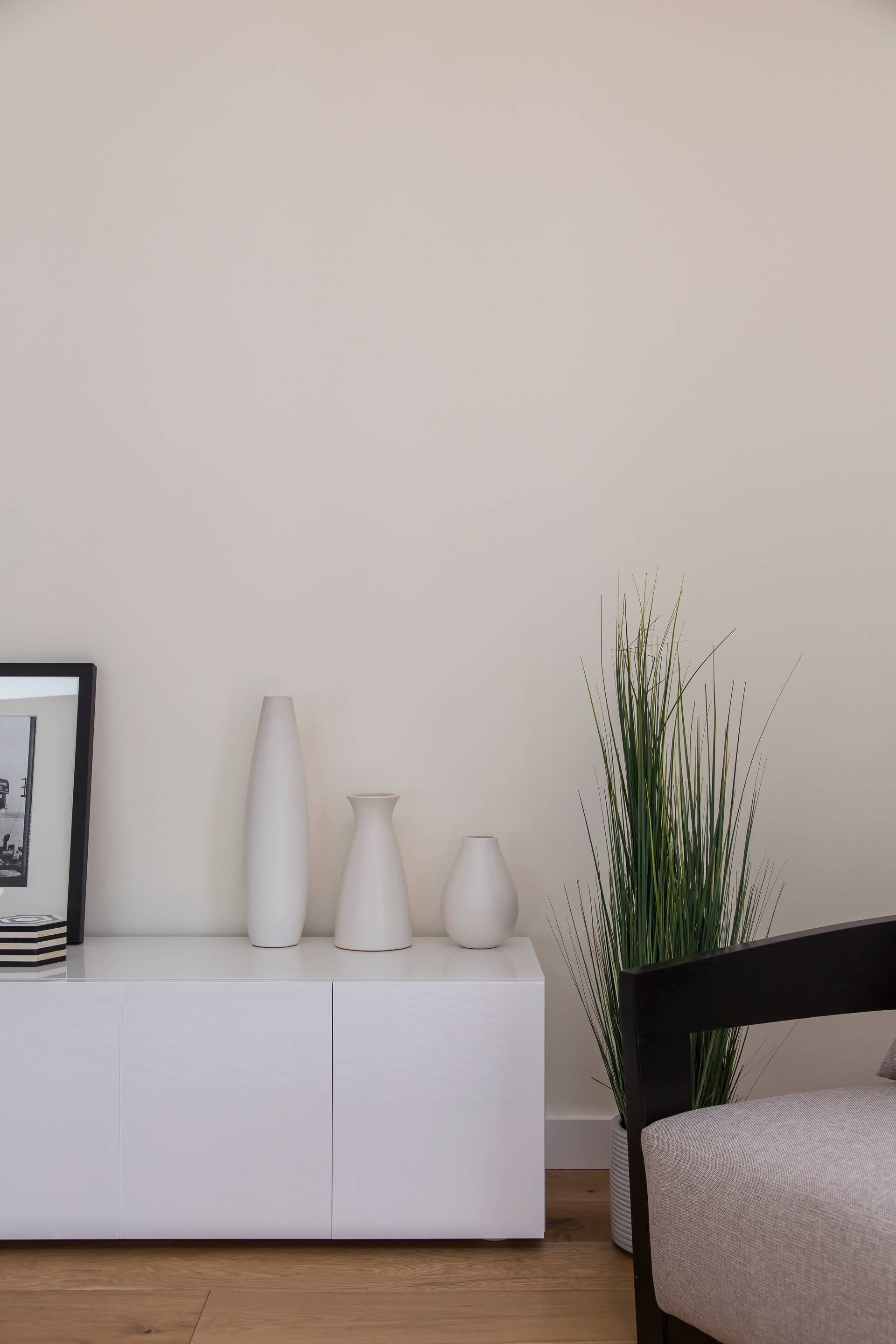 Een minimalistisch ingerichte kamer met een witte console tafel, drie witte vazen, een hoge groene plant en een lijst met een afbeelding.