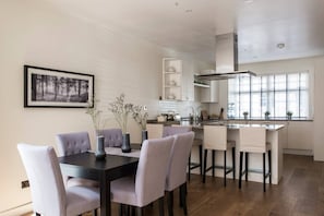 A modern kitchen with a dining area, featuring a black dining table, grey upholstered chairs, and a kitchen island with a sink and stove. - Campden Street VI by onefinestay (London)