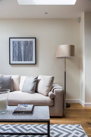 A modern living room with a sofa, a floor lamp, a framed picture on the wall, and a coffee table with books and a glass. - Campden Street VI by onefinestay (London)