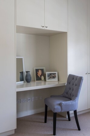A room with a white desk, a grey chair, and a shelf with a vase, a framed photo, and a picture frame.