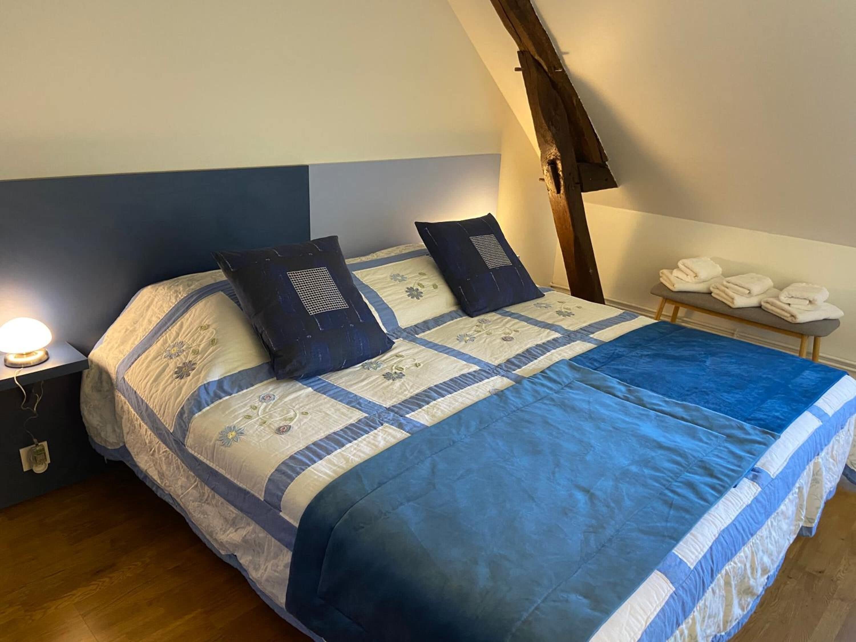 A bedroom with a bed, pillows, a bedside lamp, and a bench with towels.