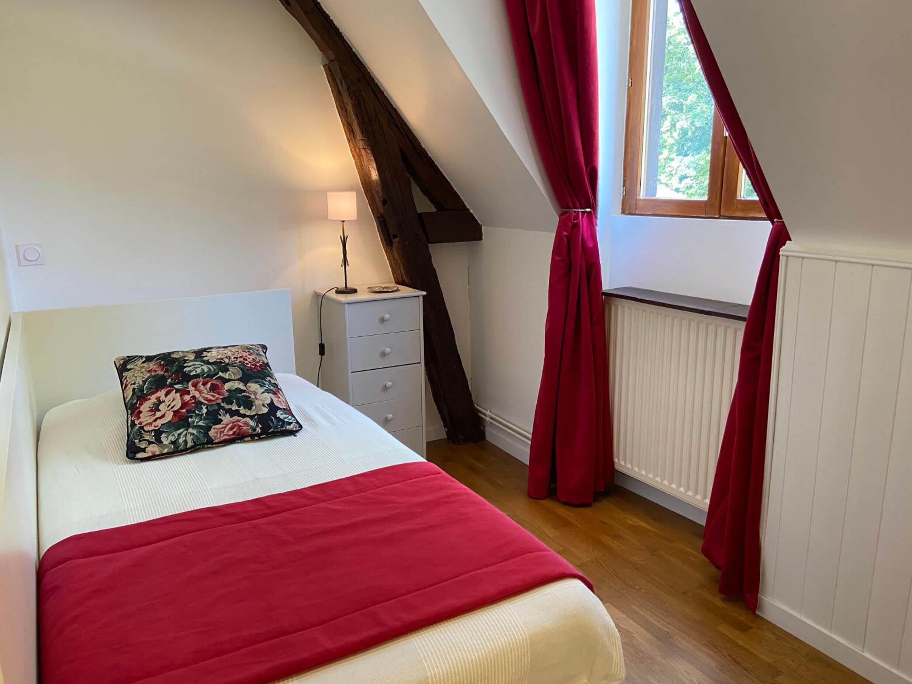 A bedroom with a bed, a nightstand, a lamp, a window with curtains, and a radiator.
