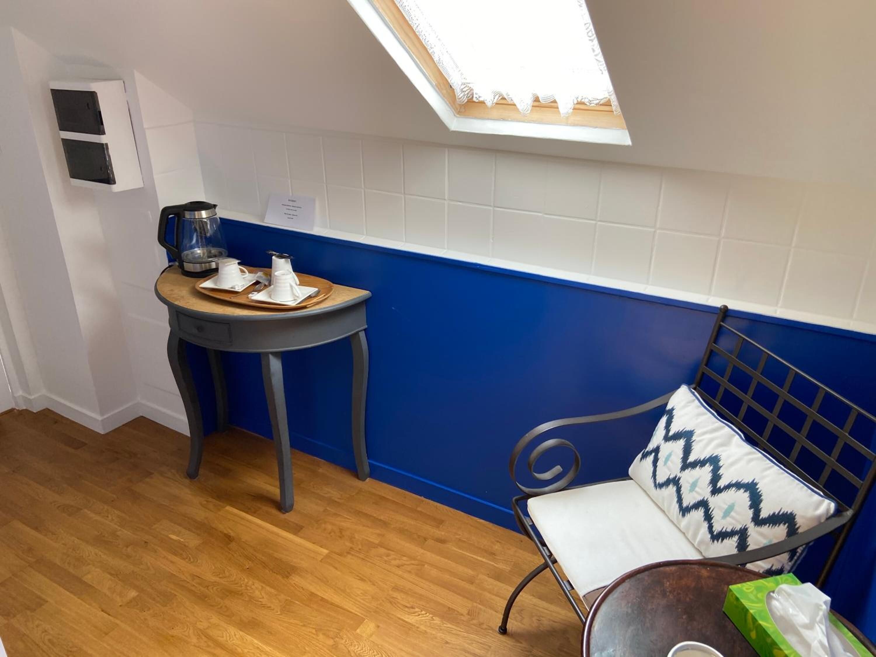 Una habitación pequeña con un tragaluz, una mesa con tazas y un hervidor, un banco con un cojín estampado, y piso de madera.