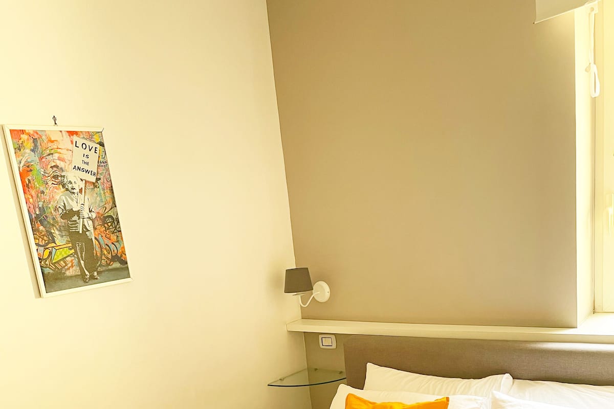 A hotel room with a bed, pillows, a glass shelf, a wall-mounted lamp, and a framed picture.