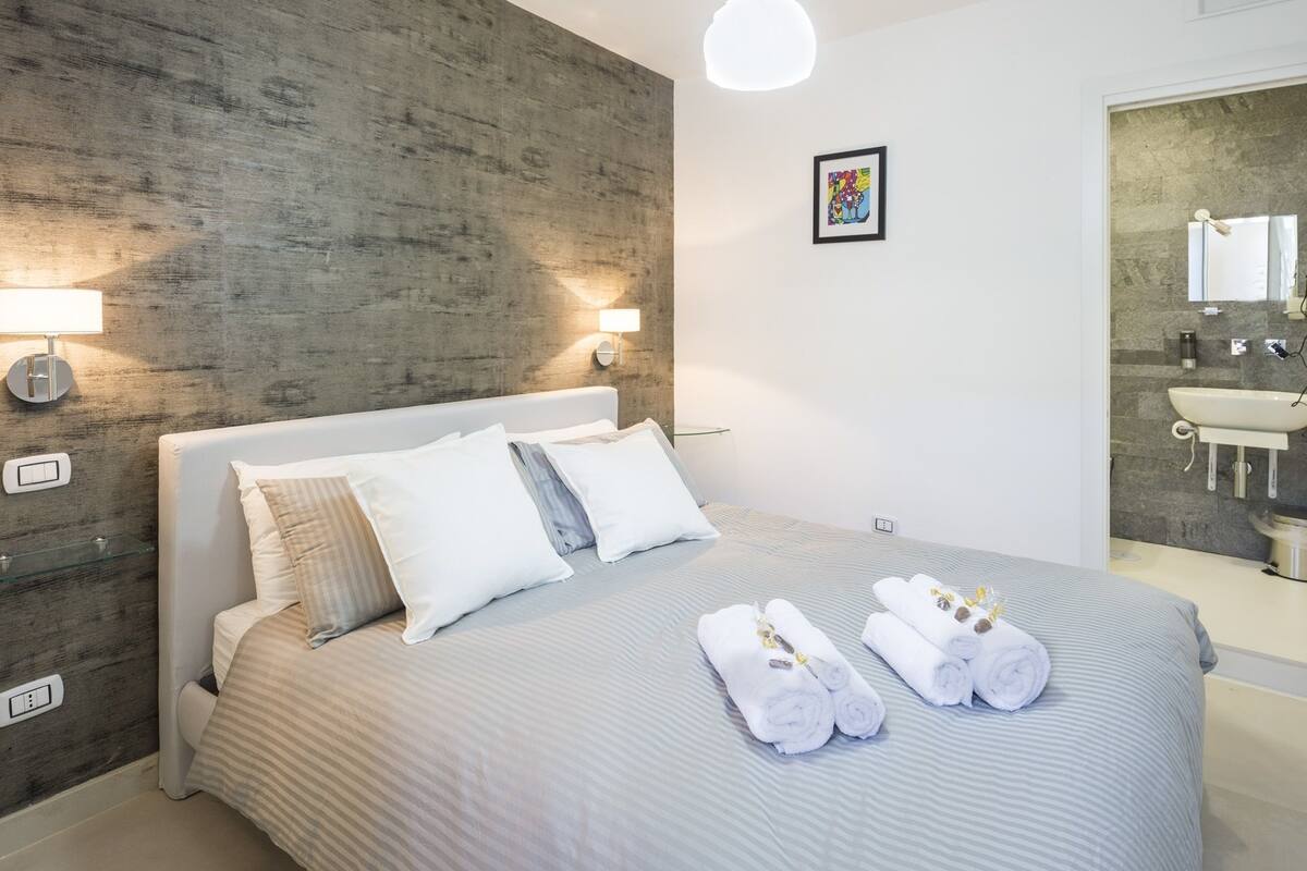 A modern hotel room with a large bed, two wall-mounted lights, a framed artwork, and a bathroom with a sink and mirror.