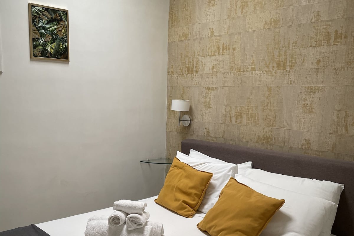A neatly made bed with white and mustard pillows, a black throw, and rolled towels.
