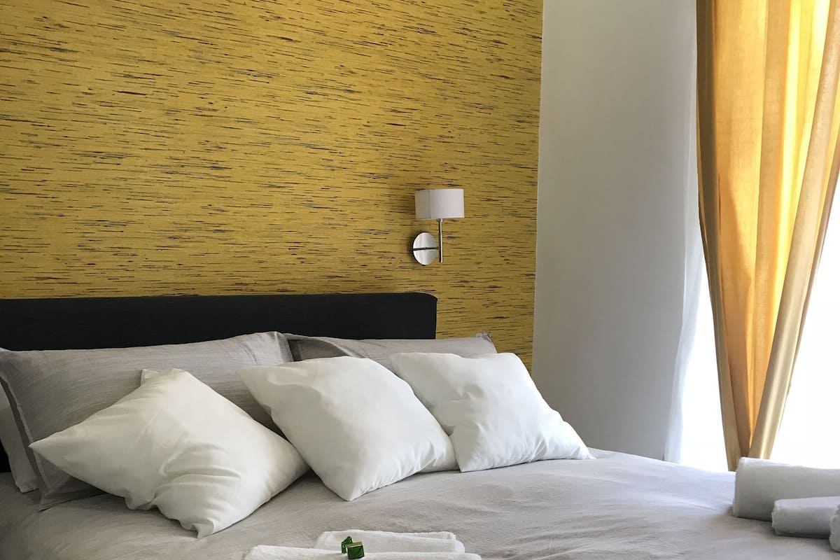 A neatly made bed with white linens and pillows, a wall-mounted lamp, and a window with curtains.