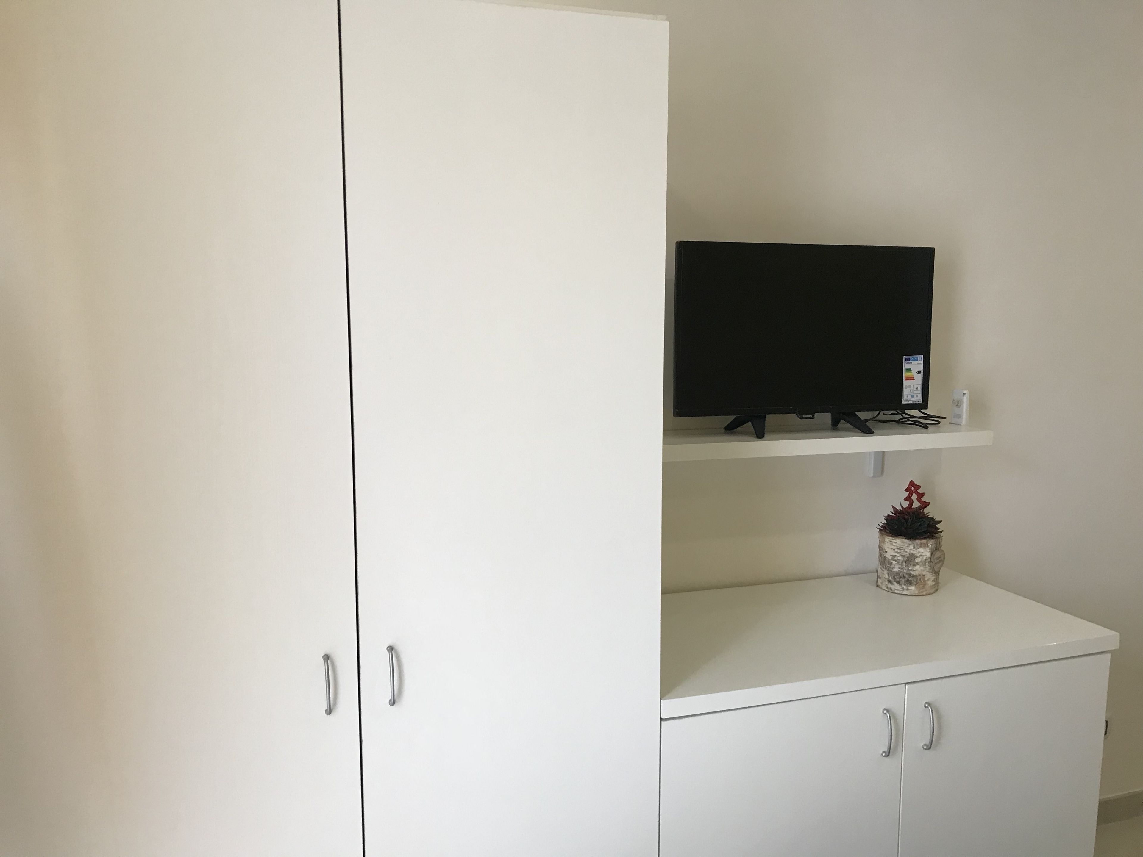 A white wardrobe with silver handles, a flat-screen TV on a white shelf, and a small potted plant.