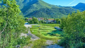 Un parcours de golf avec une chaîne de montagnes en arrière-plan, entouré d’une végétation luxuriante et de quelques bâtiments.