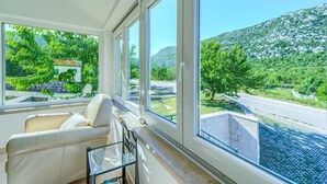 Une pièce avec un canapé et une table en verre, de grandes fenêtres donnant sur une route et sur la verdure.
