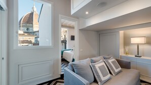 Ein modernes Hotelzimmer mit großem Fenster und Blick auf ein historisches Gebäude. Das Zimmer bietet ein bequemes Sofa, einen Nachttisch mit Lampe und ein ordentlich bezogenes Bett.