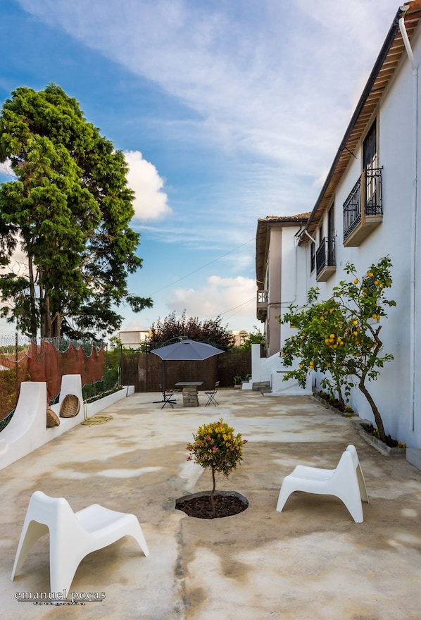 Un patio con un muro blanco, un árbol, un pequeño jardín y una silla de plástico blanca.