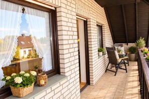 Een balkon met een bakstenen muur, een rieten mand met bloemen, een rieten stoel en potplanten.