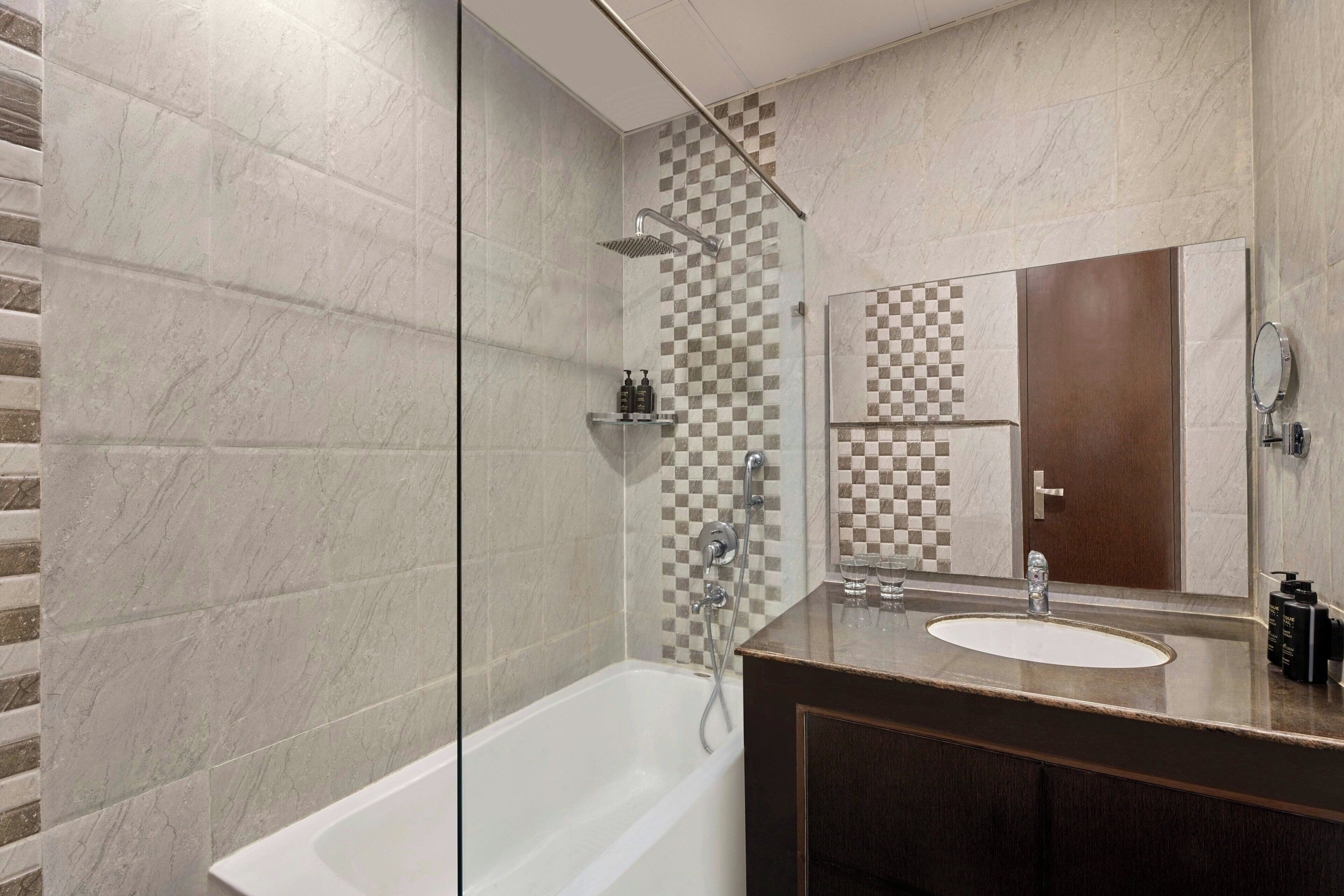 Un baño moderno con ducha de vidrio, una bañera blanca, tocador con espejo y salpicadero de baldosas a cuadros.