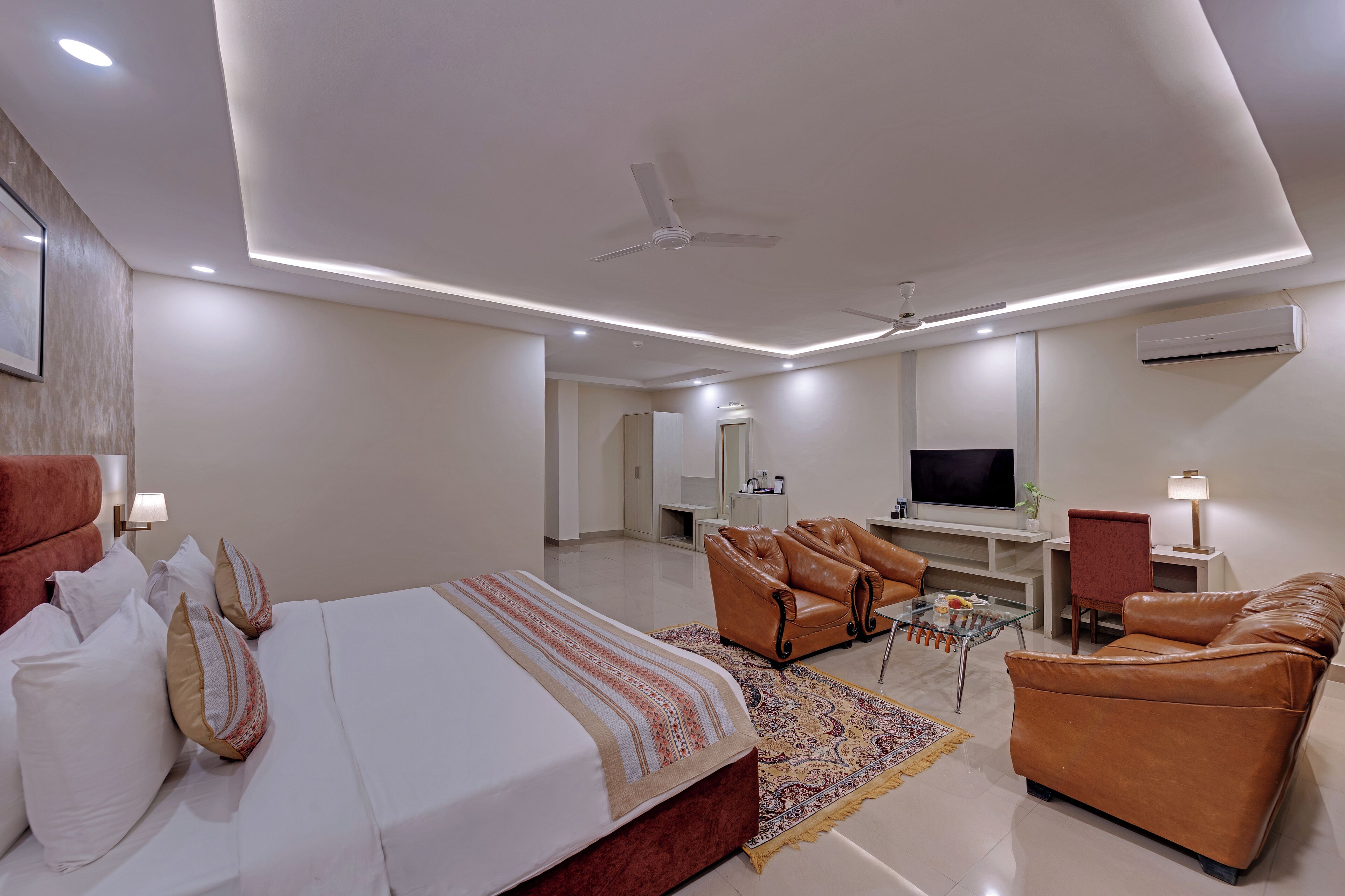 A modern hotel room with a large bed, leather sofas, a TV, and a ceiling fan.
