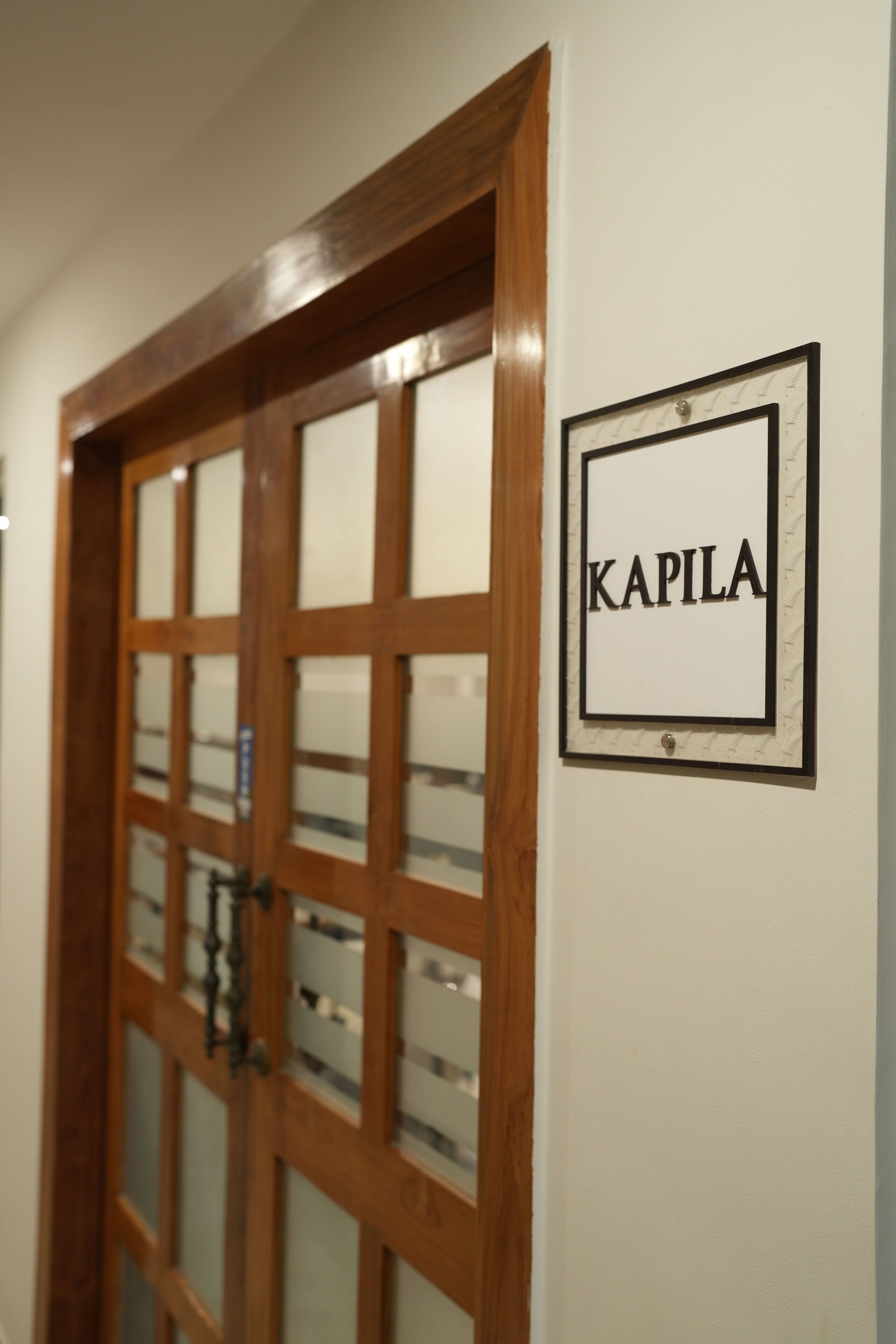 A wooden door with frosted glass panels and a sign that reads "KAPILA" on the wall.