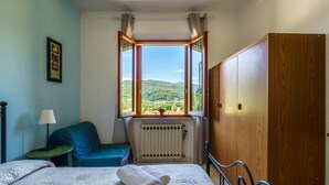 A bedroom with a bed, a wooden wardrobe, a window with a view of greenery, a radiator, and a small table with a lamp. - Residence La Mezzaluna (Barga)