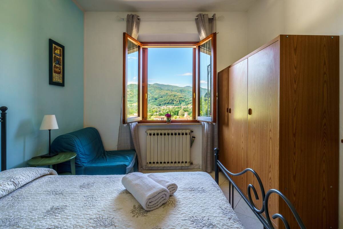 A bedroom with a bed, a wooden wardrobe, a window with a view of greenery, a radiator, and a small table with a lamp.