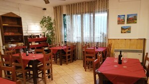 A dining area with multiple tables covered with red tablecloths, wooden chairs, and wine glasses. There is a large window with curtains, a plant, and wall art. - Residence La Mezzaluna (Barga)