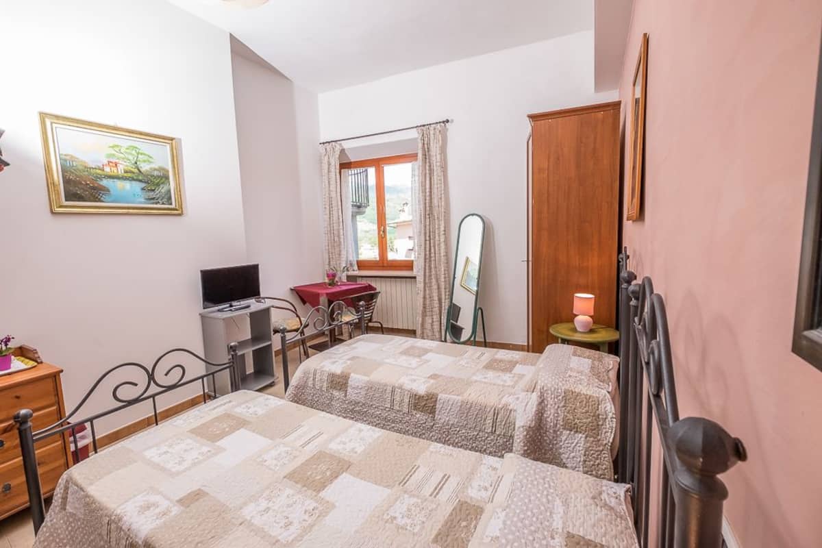 A bedroom with two beds, a desk, a chair, a TV, a window with curtains, a mirror, and a lamp.