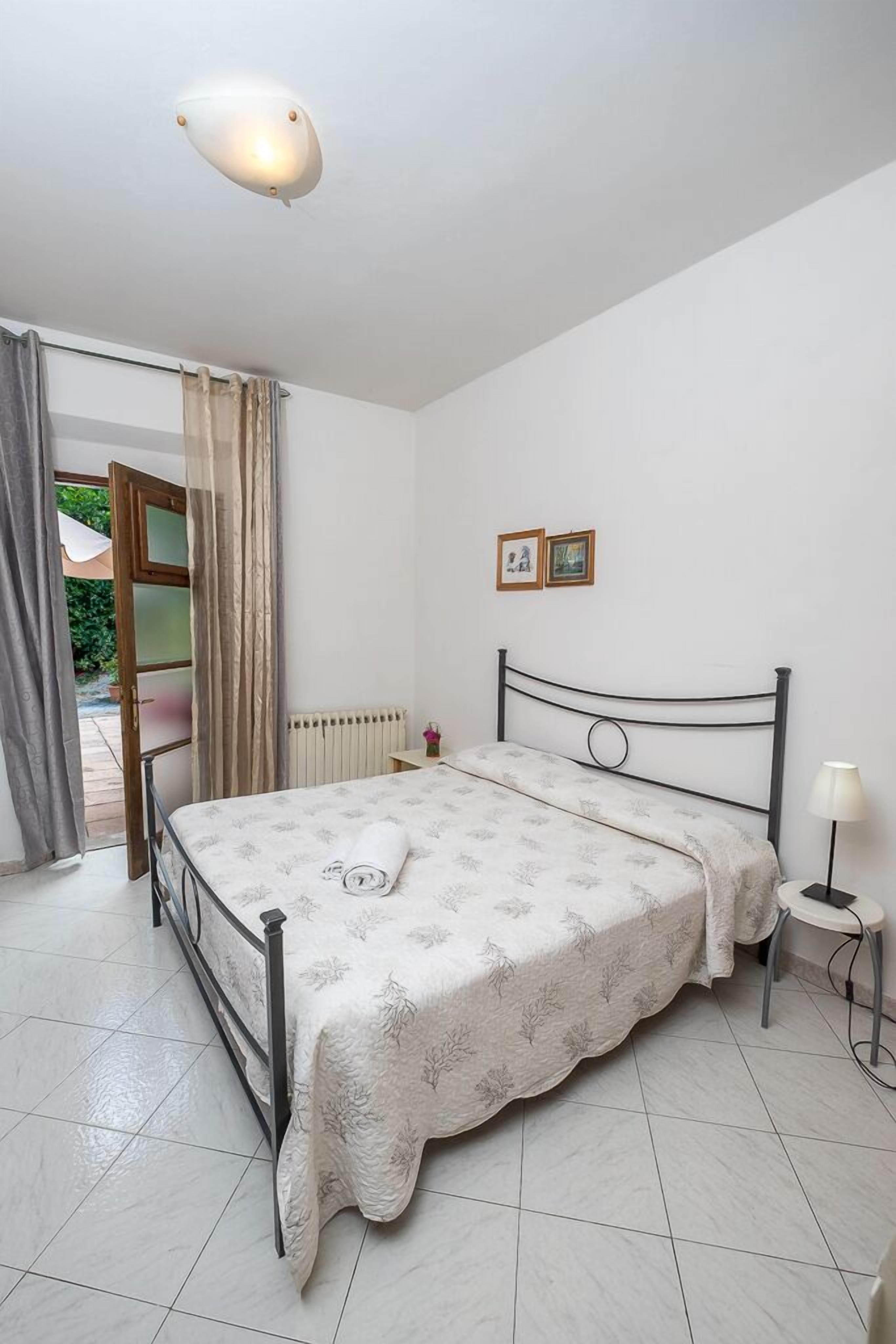 A bedroom with a bed, a nightstand, a lamp, a radiator, and a door leading to an outdoor area.