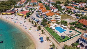 An aerial view of a coastal resort with a sandy beach, a swimming pool, and multiple buildings with red-tiled roofs. - Beautiful apartment in Povljana (Povljana)
