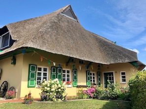 A building with a thatched roof, green shutters, and a small garden with flowers and a birdcage.