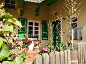 A house with green shutters, a small garden with flowers, a windmill model, and a wooden fence.
