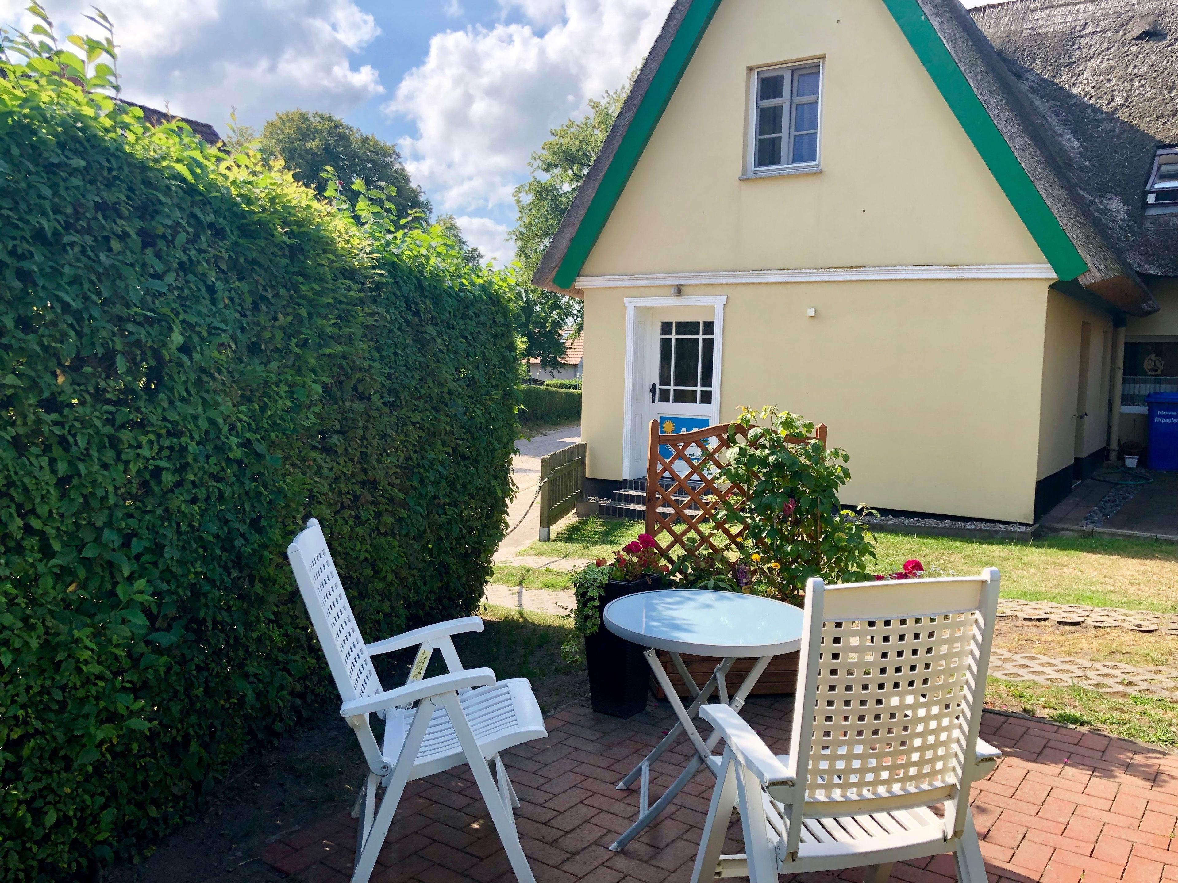 Une maison avec un toit végétalisé, une porte blanche et une petite fenêtre. On aperçoit une chaise blanche et une table au premier plan.