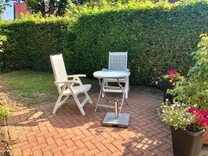 A patio with two white chairs and a table, surrounded by a hedge and a brick floor.
