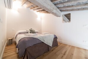 A bedroom with a wooden ceiling, a bed with a grey and white duvet, and a small window.