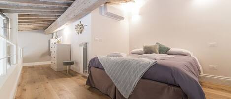 A bedroom with a wooden ceiling, a bed with a purple bedspread, a small bedside table, and a radiator.